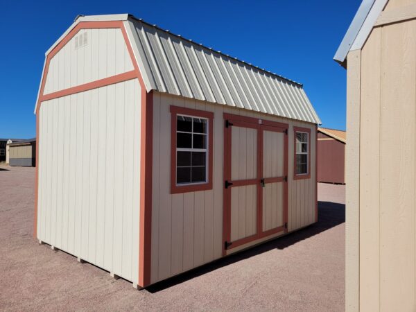 Barn style storage shed with double doors and two windows. Tan metal roof and walls with red trim.