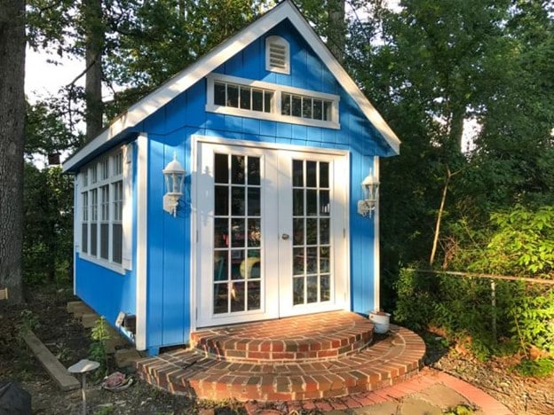 A blue gable style storage shed with brick laid in front, nestled in the forest.