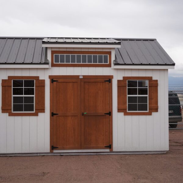 A 12x16 Studio Gable metal roofed storage shed with wooden doors and window trim, surrounded by a cloud-filled sky and several vehicles in the background.
