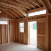 A wooden 12x16 Studio Gable storage shed is being constructed with hardwood beams, planks, plywood, swinging doors, and windows letting natural light into the building.