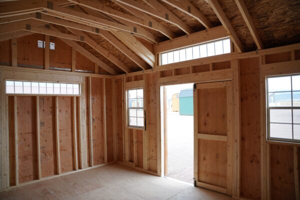 A wooden 12x16 Studio Gable storage shed is being constructed with hardwood beams, planks, plywood, swinging doors, and windows letting natural light into the building.