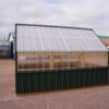 Side view of an 8x12 Green House with wood frame, green metal siding and roof, and plastic covering on the upper walls.