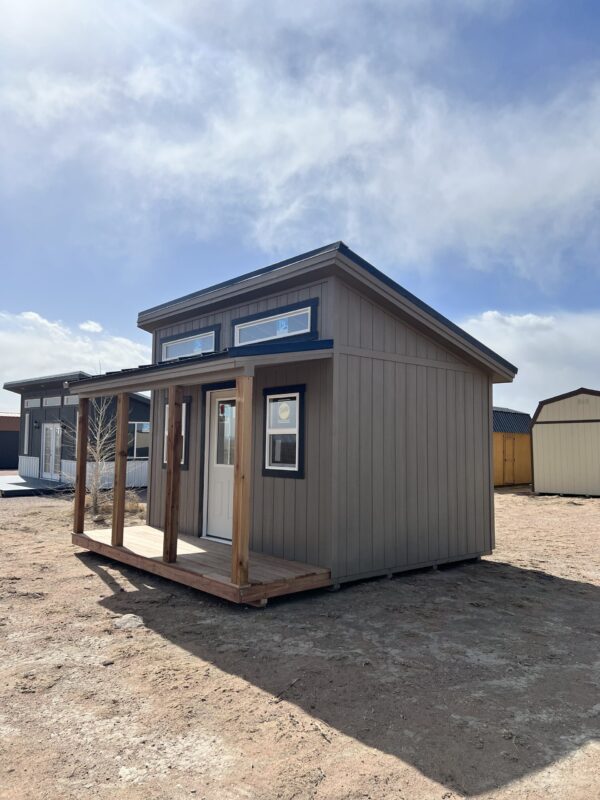 Welcome guests in style with our 14x14 Outpost Style storage shed, featuring a front view that highlights its abundance of windows, inviting wooden porch, charming wooden paneling, and timeless beige color.