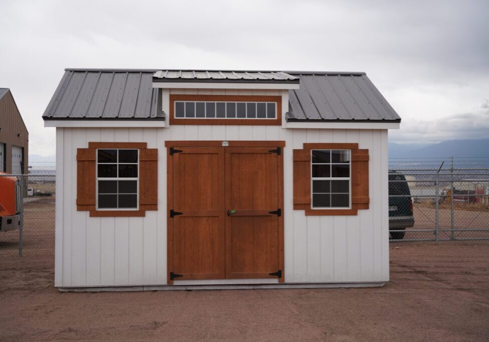 A 12x16 Studio Gable metal roofed storage shed with wooden doors and window trim, surrounded by a cloud-filled sky and several vehicles in the background.