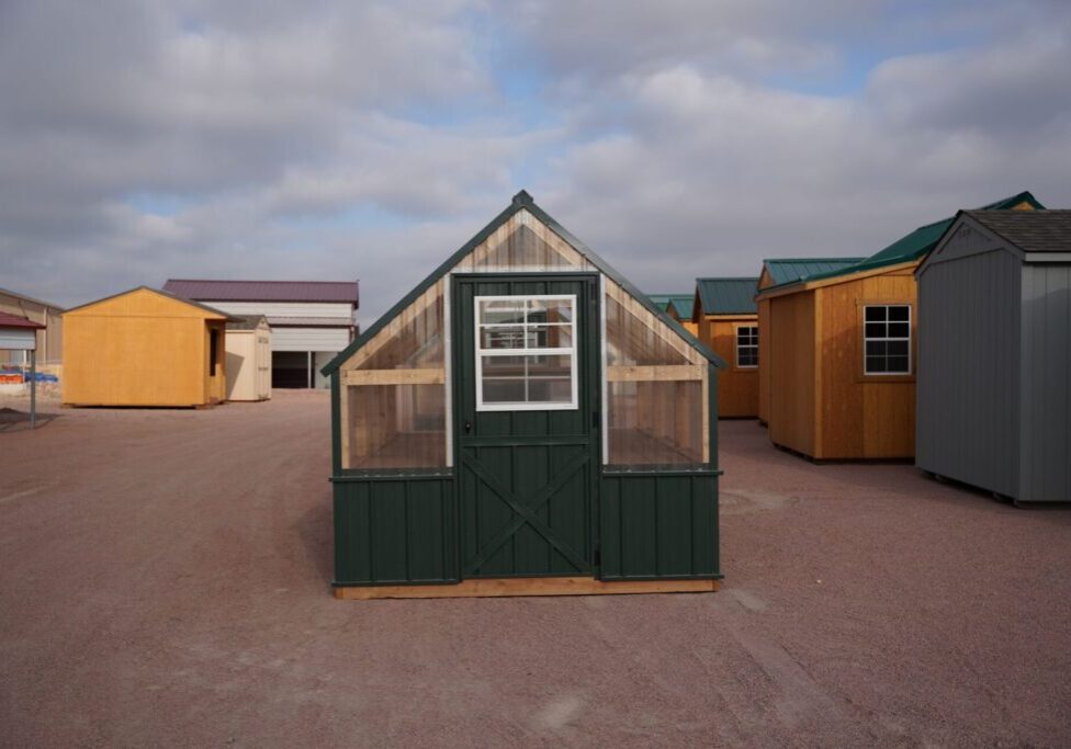 Front view of an 8x12 Green House with wood frame, green metal siding and roof, and plastic covering on the upper walls, highlighting the door in the front.